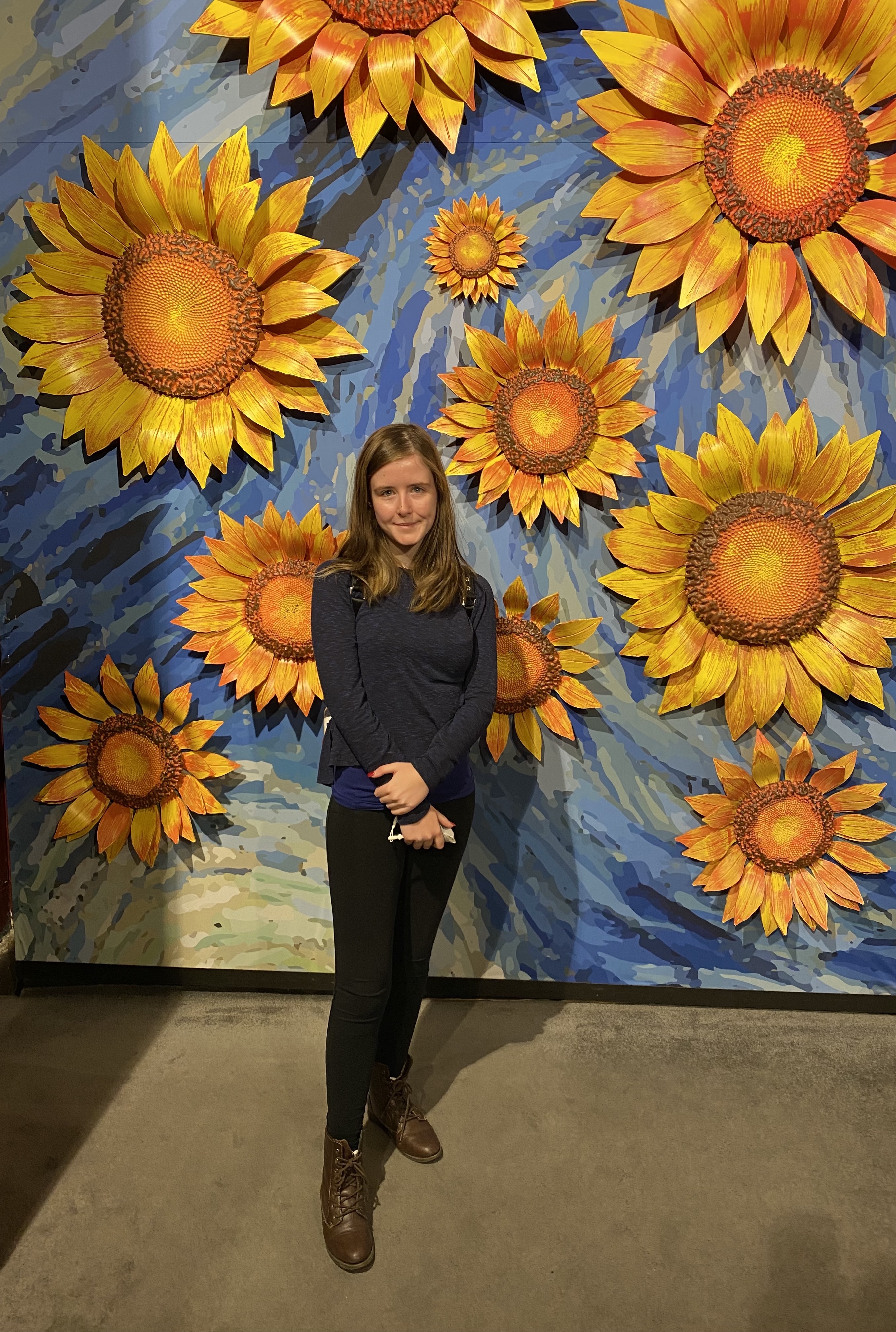 A picture of me: a girl with dark brown hair in a blue sweater, black leggings and short brown boots standing in front of a wall with 3D yellow sunflowers on a blue swirling background
